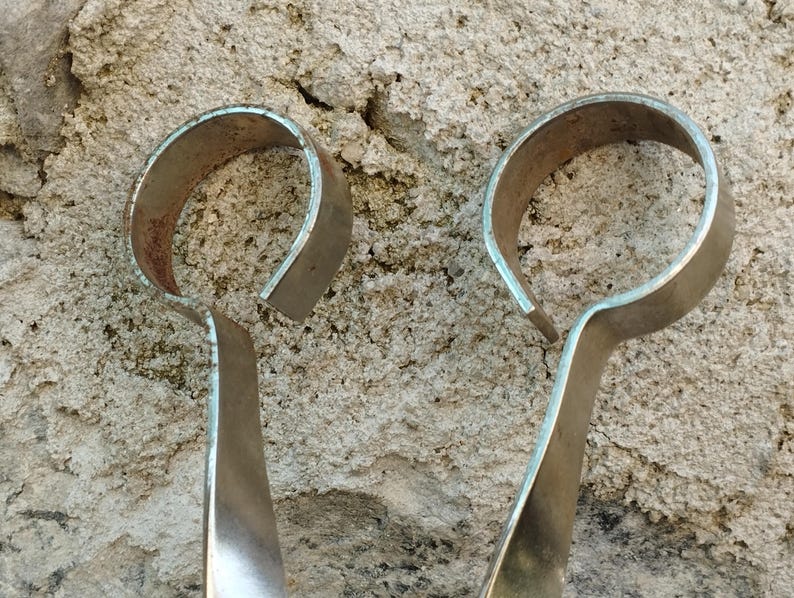 May include: A pair of silver metal tongs with rounded handles. The handles are bent into a U-shape.