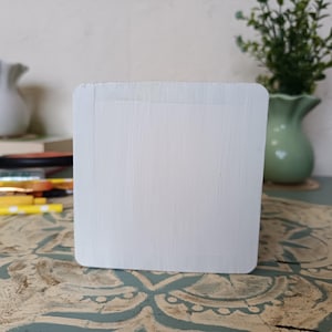 Puede incluir: Una caja de madera blanca con una superficie lisa y pintada. La caja es cuadrada y tiene un borde ligeramente redondeado. La caja est&aacute; sentada sobre una superficie estampada.