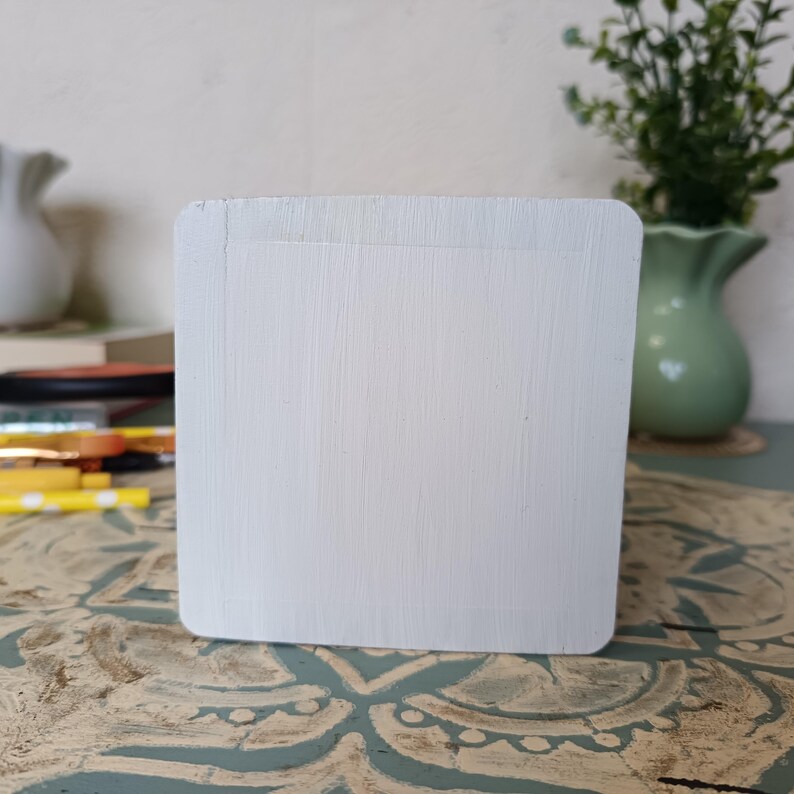 Puede incluir: Una caja de madera blanca con una superficie lisa y pintada. La caja es cuadrada y tiene un borde ligeramente redondeado. La caja est&aacute; sentada sobre una superficie estampada.