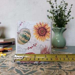 Puede incluir: Un macetero cuadrado blanco con un dise&ntilde;o floral que presenta un girasol, una calabaza y bayas rojas. El macetero tiene 7,6 cm de alto y se est&aacute; midiendo con una cinta m&eacute;trica.