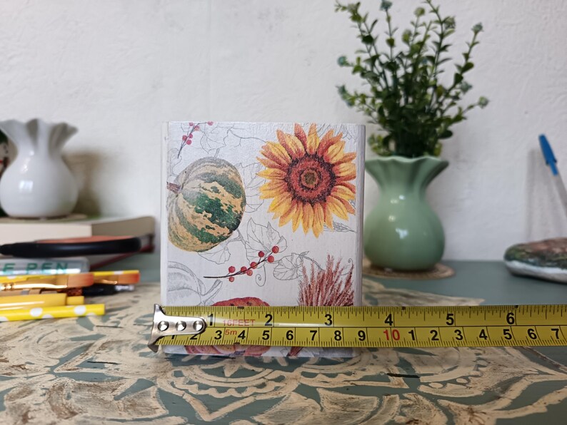 Puede incluir: Un macetero cuadrado blanco con un dise&ntilde;o floral que presenta un girasol, una calabaza y bayas rojas. El macetero tiene 7,6 cm de alto y se est&aacute; midiendo con una cinta m&eacute;trica.