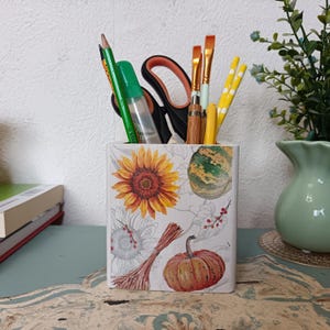 Puede incluir: Un porta l&aacute;pices de cer&aacute;mica blanca con un dise&ntilde;o de oto&ntilde;o con girasoles, calabazas y calabacines. El porta l&aacute;pices est&aacute; lleno de l&aacute;pices, bol&iacute;grafos y pinceles.