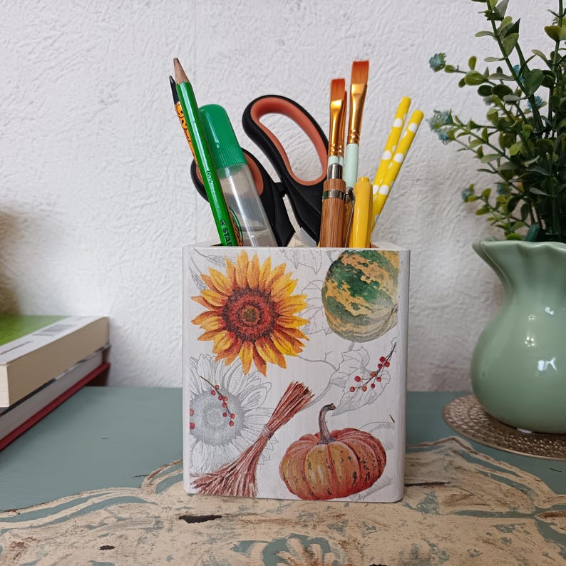 Puede incluir: Un porta l&aacute;pices de cer&aacute;mica blanca con un dise&ntilde;o de oto&ntilde;o con girasoles, calabazas y calabacines. El porta l&aacute;pices est&aacute; lleno de l&aacute;pices, bol&iacute;grafos y pinceles.