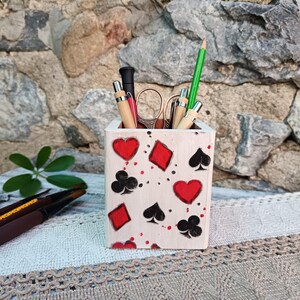 May include: A white wooden desk organizer decorated with playing card suits: hearts, diamonds, clubs, and spades. The organizer holds pens, pencils, and scissors. The background features a stone wall and a decorative lace tablecloth.