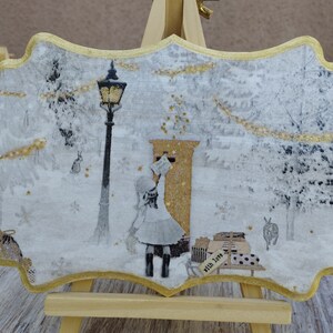 May include: A white and gold decorative plaque with a winter scene. A girl in a white dress is standing in front of a gold mailbox. The scene is surrounded by a gold frame.