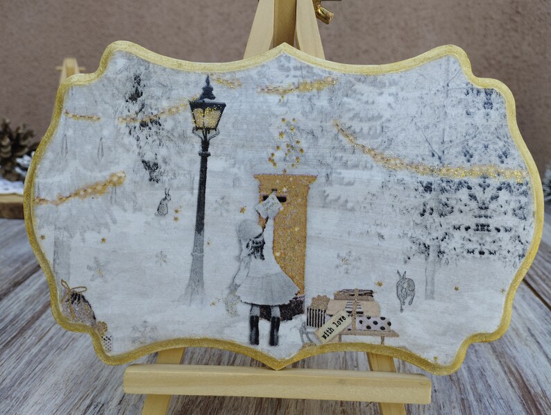 May include: A white and gold decorative plaque with a winter scene. A girl in a white dress is standing in front of a gold mailbox. The scene is surrounded by a gold frame.