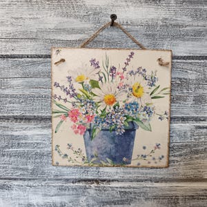 Op de afbeelding: Een vierkant houten bord met een geschilderd beeld van een blauwe emmer gevuld met een boeket witte margrieten, paarse lavendel en blauwe vergeet-mij-nietjes.