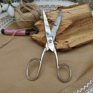 May include: A pair of silver-toned scissors with a wooden handle. The scissors are open and the blades are sharp. The scissors are on a brown surface.
