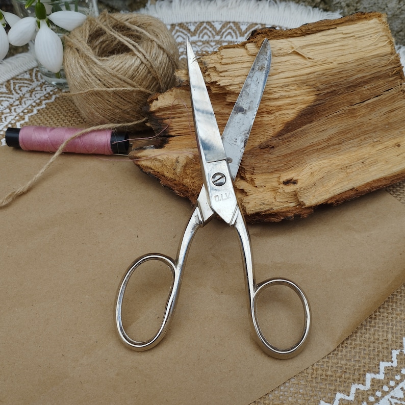 May include: A pair of silver-toned scissors with a wooden handle. The scissors are open and the blades are sharp. The scissors are on a brown surface.