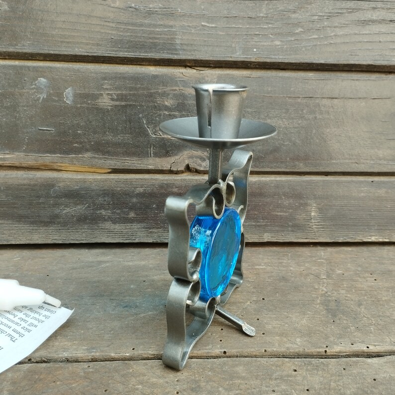 Pu&ograve; includere: Un portacandele in metallo con finitura grigio opaco e volute decorative che incorniciano un inserto circolare in vetro blu traslucido. La struttura include una coppa cilindrica per candele in alto e un piccolo piedino di supporto in metallo.