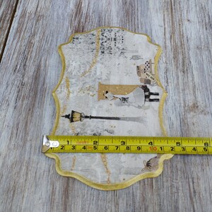 May include: A decorative, gold-trimmed, white paper plaque with a winter scene featuring a woman in a white dress and a street lamp. The plaque is approximately 7 inches wide.