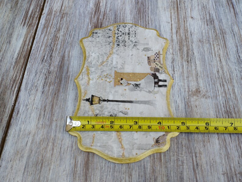 May include: A decorative, gold-trimmed, white paper plaque with a winter scene featuring a woman in a white dress and a street lamp. The plaque is approximately 7 inches wide.