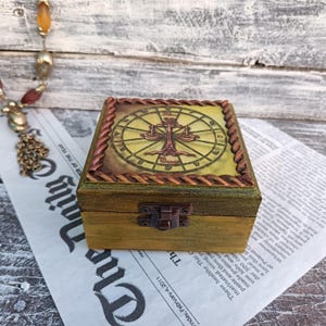 May include: A wooden box with a hinged lid and a brass latch. The top of the box is decorated with a painted image of the Libra zodiac sign, surrounded by a rope-like border. The background is a light green colour.