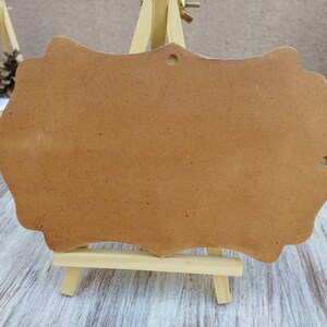 May include: A brown wooden sign with a scalloped edge, standing on a small easel. The sign is blank and ready to be decorated.