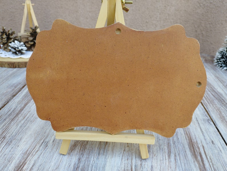 May include: A brown wooden sign with a scalloped edge, standing on a small easel. The sign is blank and ready to be decorated.