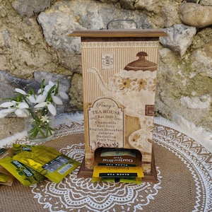 Könnte beinhalten: Eine Holzteekiste mit einem Vintage-Design mit einer Teekanne und Blumen. Die Kiste hat einen Spender für Teebeutel und ist mit "Penny's Tea House Est. 1890" beschriftet. Die Kiste ist mit Teebeuteln in verschiedenen Geschmacksrichtungen gefüllt, darunter schwarzer Tee, englisches Frühstück und Darjeeling.