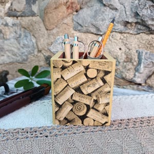 May include: A wooden desk organizer with a cork design, holding pens, pencils, and a pair of glasses. The organizer is rectangular with a light brown exterior and a red interior. The cork design features various wine corks with printed text.