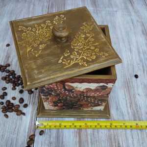 May include: A wooden box with a hinged lid decorated with a floral design. The box is painted in a light brown color and has a brown knob on the lid. The inside of the box is lined with a brown and white patterned paper. The box is sitting on a wooden surface.