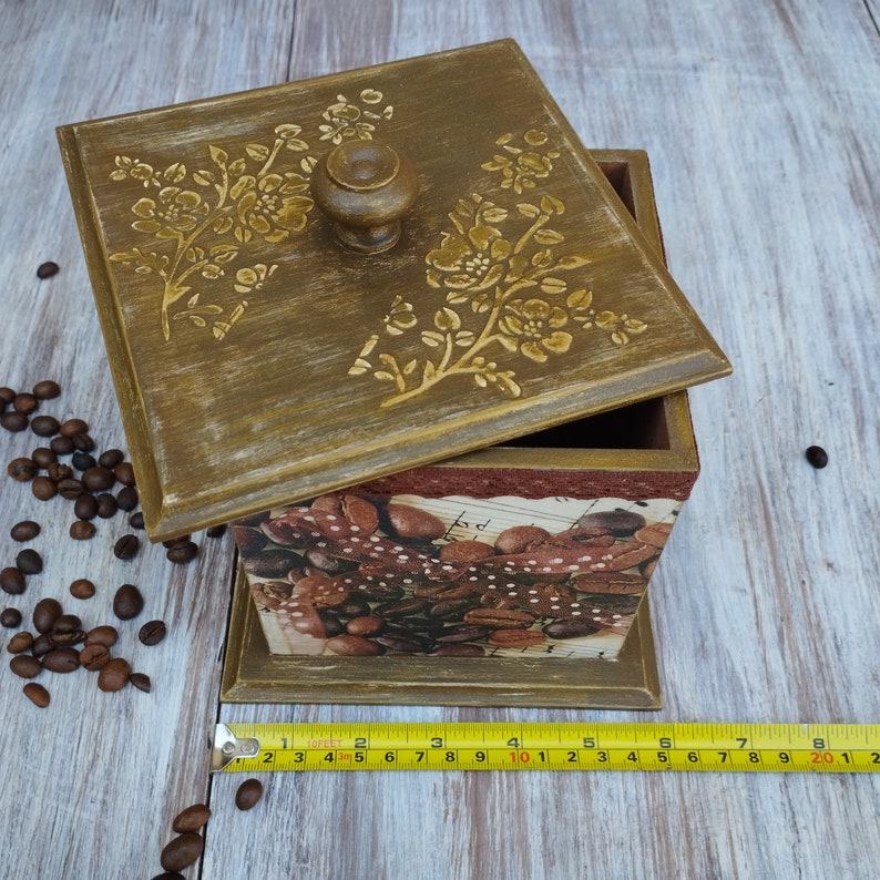 May include: A wooden box with a hinged lid decorated with a floral design. The box is painted in a light brown color and has a brown knob on the lid. The inside of the box is lined with a brown and white patterned paper. The box is sitting on a wooden surface.