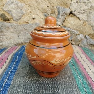 Pode incluir: Um pequeno pote de cerâmica marrom com tampa. O pote tem um design decorativo de linhas pretas e brancas e folhas marrons. A tampa tem uma linha ondulada branca ao redor da parte superior.