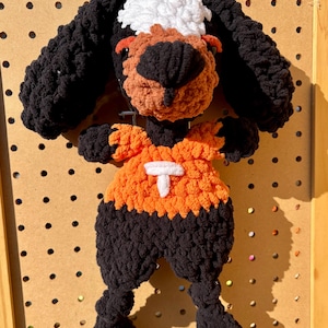 May include: A handmade crochet dog toy with a black body, ears, and legs. The dog's face is brown and black with a white patch on top of its head. It wears an orange shirt with a white "T" on the front.