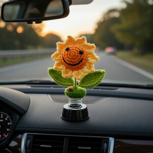 May include: A crocheted sunflower car accessory with a smiling face, yellow petals, and green leaves. The flower sits on a black base with a spring, adding a playful touch to the car's dashboard. A small red ladybug is on the green base.