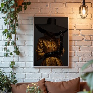 Op de afbeelding: Een ingelijste print van een vrouw die een grote zwarte hoed en een gouden geprinte jurk draagt. De vrouw staat voor een donkere achtergrond.