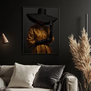 Op de afbeelding: Een zwart en gouden ingelijste print van een vrouw die een brede zwarte hoed en een gouden geprinte jurk draagt.