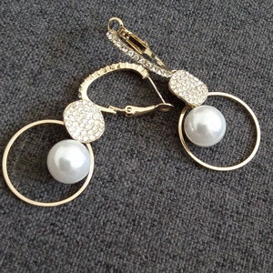 May include: A pair of gold earrings with a pearl and crystal accents. Each earring features a round hoop with a smaller round crystal-encrusted disc and a pearl hanging from the hoop.