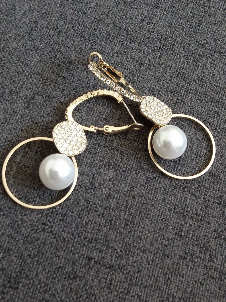 May include: A pair of gold earrings with a pearl and crystal accents. Each earring features a round hoop with a smaller round crystal-encrusted disc and a pearl hanging from the hoop.
