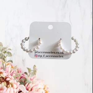 May include: A pair of white pearl hoop earrings with gold posts. The earrings are displayed on a white card with the text "pfaccessories.co.uk @p.f.accessories".