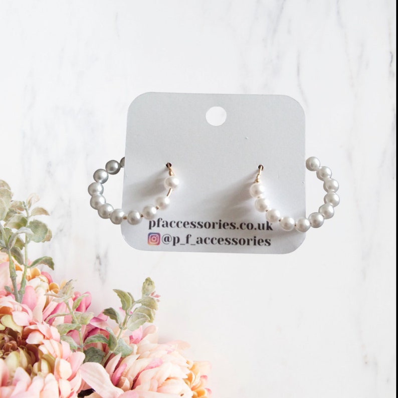 May include: A pair of white pearl hoop earrings with gold posts. The earrings are displayed on a white card with the text "pfaccessories.co.uk @p.f.accessories".