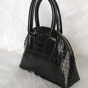 May include: A black faux leather handbag with a crocodile embossed pattern and gold-toned hardware. The bag has two top handles and a zipper closure.