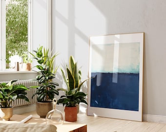 May include: A framed abstract painting with a blue and white color scheme hangs on a white wall in a living room. The painting is in a wooden frame and is surrounded by houseplants.