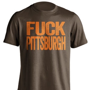 May include: A brown t-shirt with the words "FUCK PITTSBURGH" printed in large, distressed orange letters across the front. The shirt has a crew neck and short sleeves.