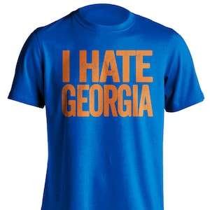 May include: Royal blue t-shirt with the orange text "I HATE GEORGIA". The shirt has a crew neck and short sleeves. The text has a distressed, vintage look.