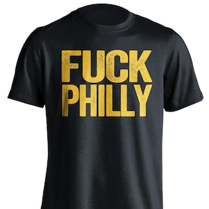 May include: Black t-shirt with the words "FUCK PHILLY" printed in distressed yellow block letters. The shirt has a crew neck and short sleeves. The text is centered on the front of the shirt.