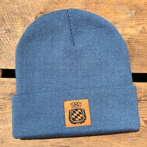 Könnte beinhalten: Eine blaue Strickmütze mit einem braunen Lederpatch mit einem schwarz-weiß karierten Muster und einer Krone.
