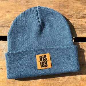 Könnte beinhalten: Eine blaue Strickmütze mit einem braunen Lederpatch, auf dem "ois-isy" in schwarzen Buchstaben steht.