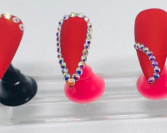 May include: Five red matte artificial nails with various rhinestone designs. The nails are displayed on a clear plastic stand with black and pink holders. Each nail has a different rhinestone pattern.