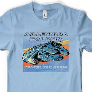 May include: A light blue t-shirt featuring a retro-style graphic of the Millennium Falcon spaceship. The graphic includes the text "Millennium Falcon" and "The fastest hunk of junk in the galaxy".