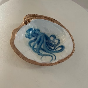 May include: A decorative seashell dish with a gold-painted rim. The interior features a blue octopus design on a white background. The dish is set on a white surface.