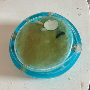 Puede incluir: Un cenicero redondo de resina con un borde azul turquesa y un centro verde. El cenicero contiene pequeñas conchas marinas y una concha blanca. La resina transparente permite ver el contenido. Un artículo decorativo.
