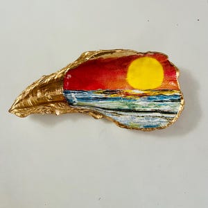 May include: A seashell painted with a sunset scene. The shell is gold and the sunset is painted in shades of red, orange, yellow, and blue. The ocean is painted in shades of blue and green.
