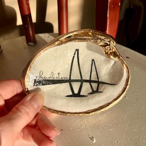 May include: A white seashell dish with a gold rim, featuring a black silhouette of the Cooper River Bridge and the word "Charleston".