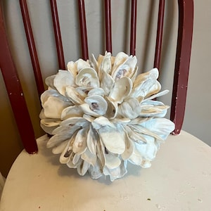 May include: A decorative sphere composed of numerous oyster shells, arranged to resemble a flower. The shells are primarily off-white with subtle brown and gray tones. The sphere rests on a white chair with a red wooden frame.