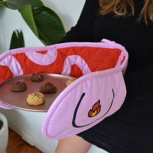 May include: Pink oven mitts with a red quilted lining and a flame design. The mitts are holding a baking sheet with cookies.