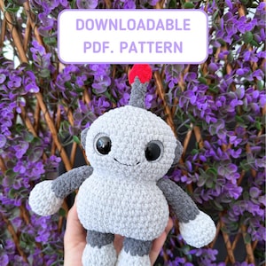 May include: A grey crocheted robot toy with large black eyes and a red pom-pom on top of its head. The toy is being held in a hand against a background of purple flowers. DOWNLOADABLE PDF. PATTERN.