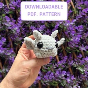 May include: A gray crocheted gargoyle amigurumi toy with black eyes. DOWNLOADABLE PDF. PATTERN is written in white text on a purple background.