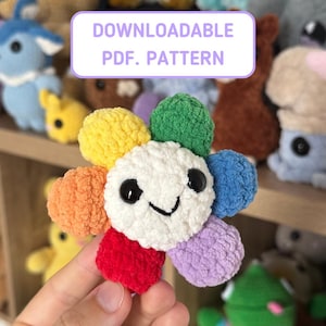 Op de afbeelding: Een kleurrijke, gehaakte bloem met een lachend gezicht en regenboogkleurige bloemblaadjes. De bloem heeft een wit hart met zwarte ogen en een gestikte glimlach. De afbeelding bevat de tekst "DOWNLOADABLE PDF. PATTERN".